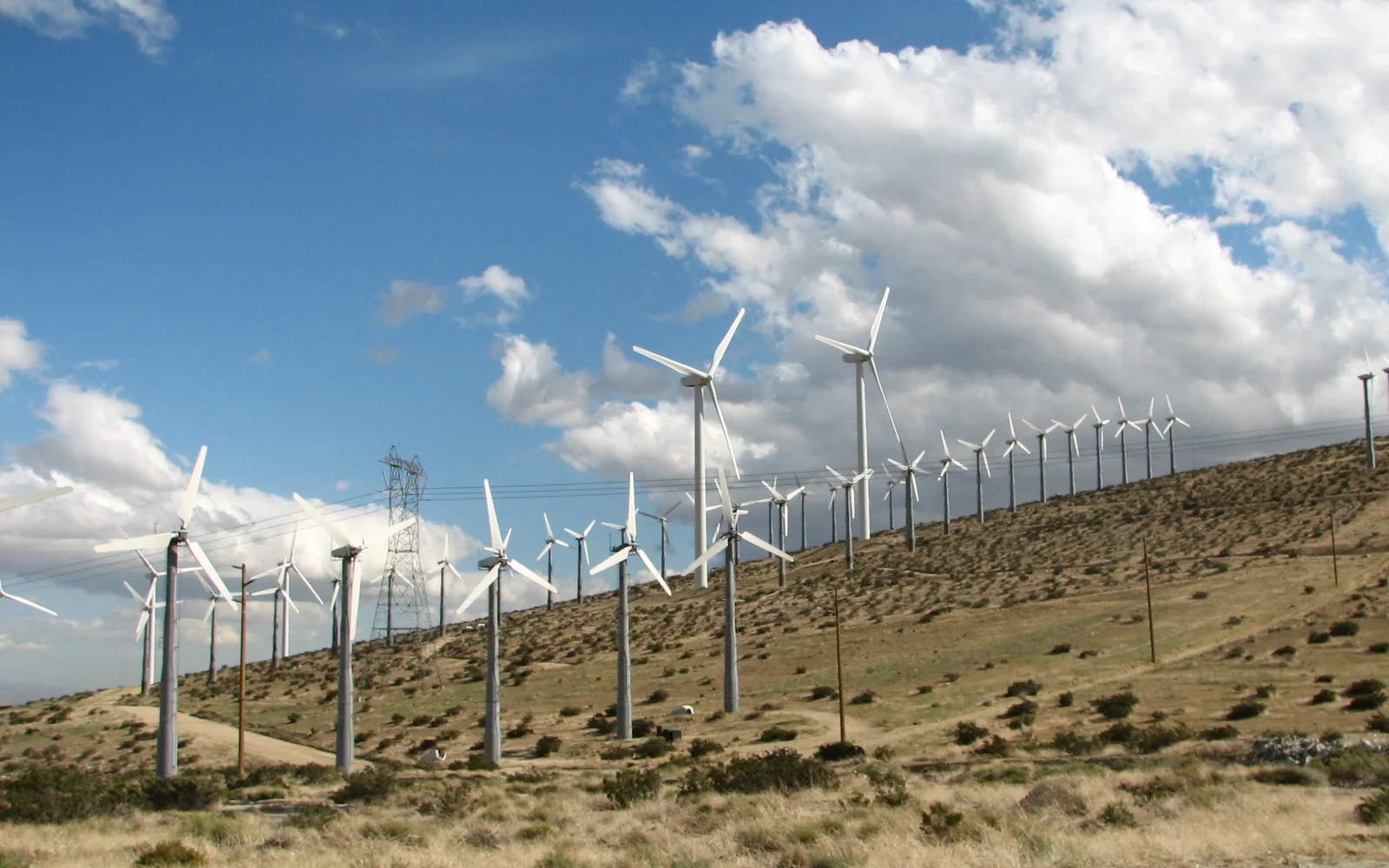 Multiple large white wind turbines in a desert landscape against a blue sky with scattered clouds